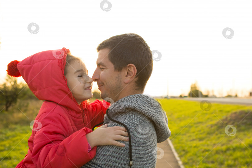 Скачать Маленькая девочка в красной куртке с капюшоном обнимает и целует своего папу, улыбается, касается своего носа. Счастливая семья, эмоции детей, день отца, яркие лучи солнца, кавказская внешность. Место для текста. фотосток Ozero
