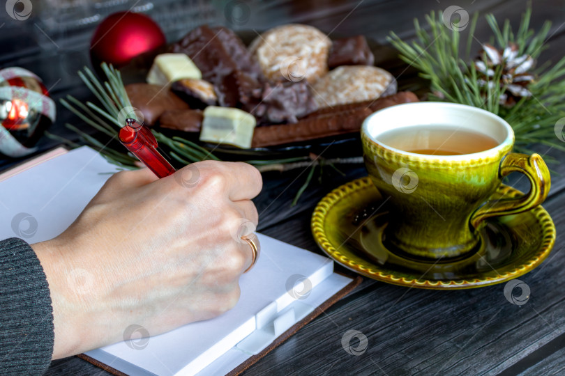 Скачать Женская рука записывает в блокнот план на новый год в уютной обстановке. Рождественский декор, сладости, чай. Хюгге фотосток Ozero