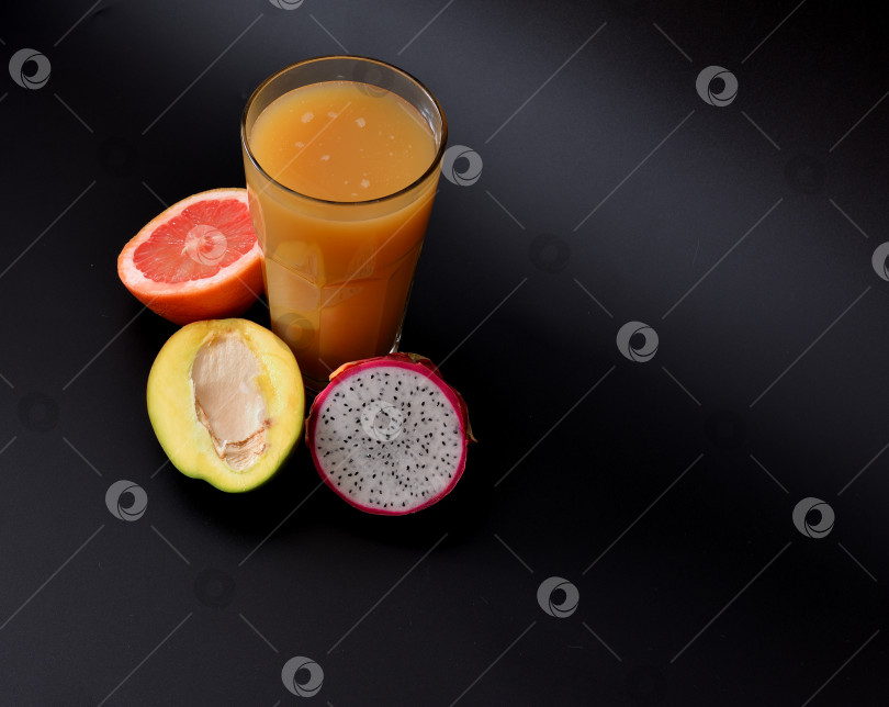 Скачать Высокий стакан свежевыжатого фруктового сока на черном фоне, рядом с половинками спелого манго, грейпфрута и питайи. фотосток Ozero