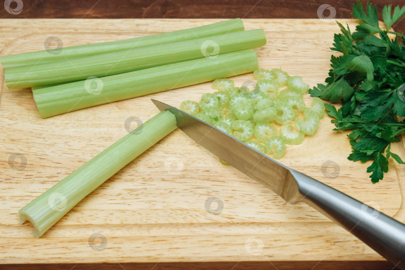 Скачать Стебли зеленого сельдерея нарезать металлическим ножом на светлой деревянной разделочной доске вместе с пучком петрушки на коричневом столе фотосток Ozero