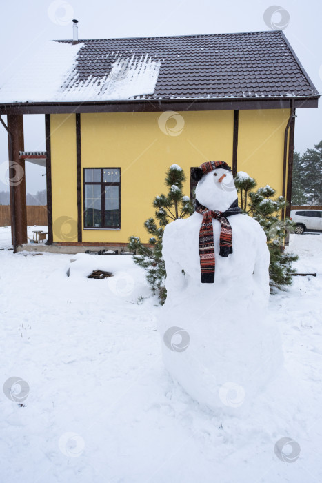 Скачать Забавный снеговик в шапке и шарфе на фоне желтого дома во дворе. Зима, зимние развлечения, снегопад фотосток Ozero