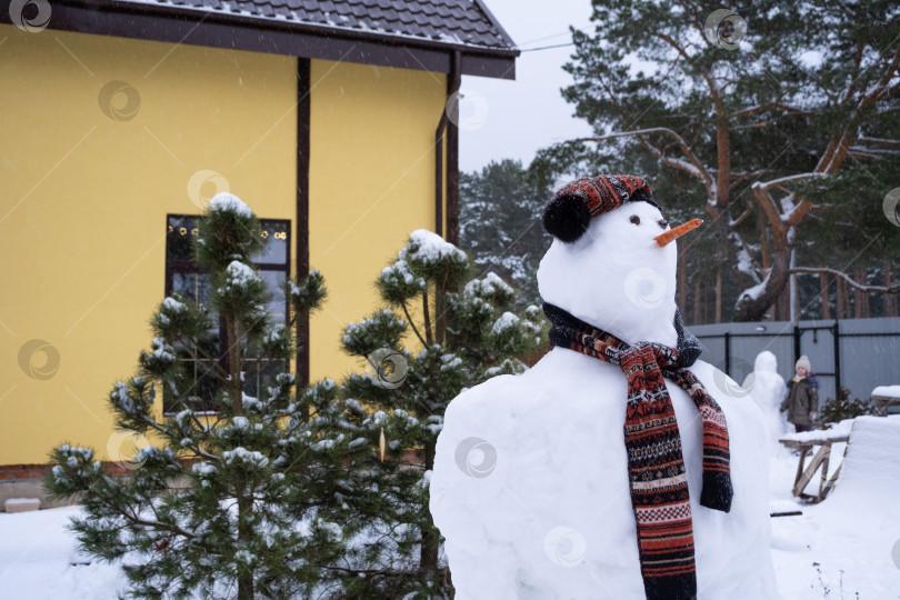 Скачать Забавный снеговик в шапке и шарфе на фоне желтого дома во дворе. Зима, зимние развлечения, снегопад фотосток Ozero