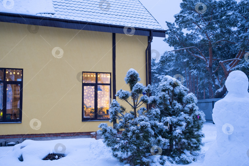 Скачать Уютное праздничное окно дома снаружи с теплым светом гирлянд fairy lights внутри - отпразднуйте Рождество и Новый год в теплом доме. Рождественская елка, боке, снег на соснах и снегопад фотосток Ozero