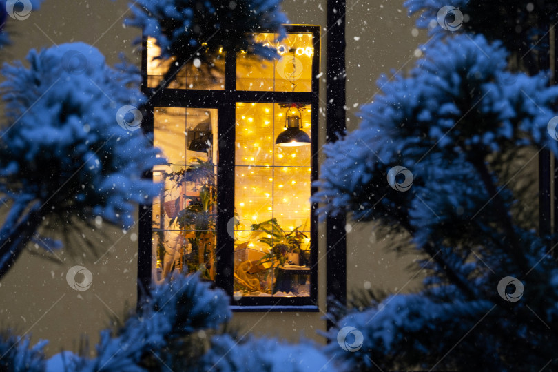 Скачать Уютное праздничное окно дома снаружи с теплым светом гирлянд fairy lights внутри - отпразднуйте Рождество и Новый год в теплом доме. Рождественская елка, боке, снег на соснах и снегопад фотосток Ozero