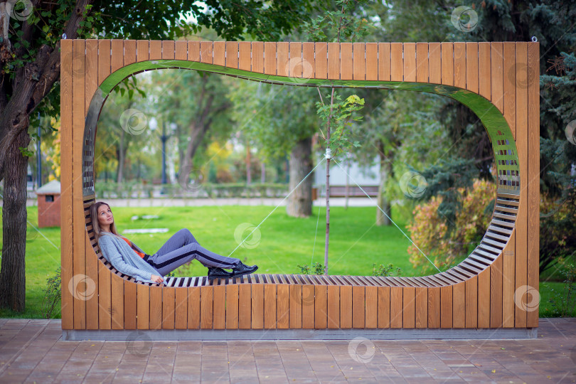 Скачать Красивая девушка в сером свитере сидит, поджав ноги, и отдыхает на круглой скамейке в парке фотосток Ozero