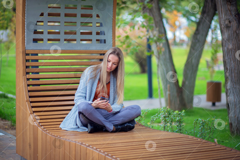 Скачать Девушка с длинными волосами и в сером свитере сидит на скамейке в парке и смотрит в смартфон фотосток Ozero