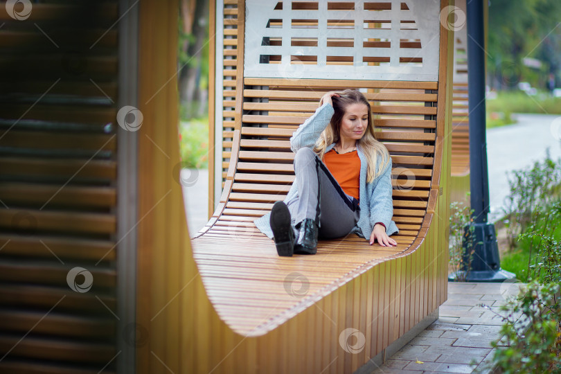 Скачать Красивая девушка сидит, поджав ноги, и отдыхает на большой современной скамейке в парке фотосток Ozero