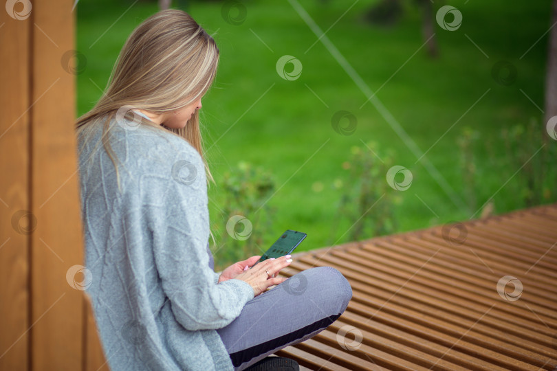 Скачать Белокурая девушка сидит с ногами на скамейке и смотрит в смартфон фотосток Ozero