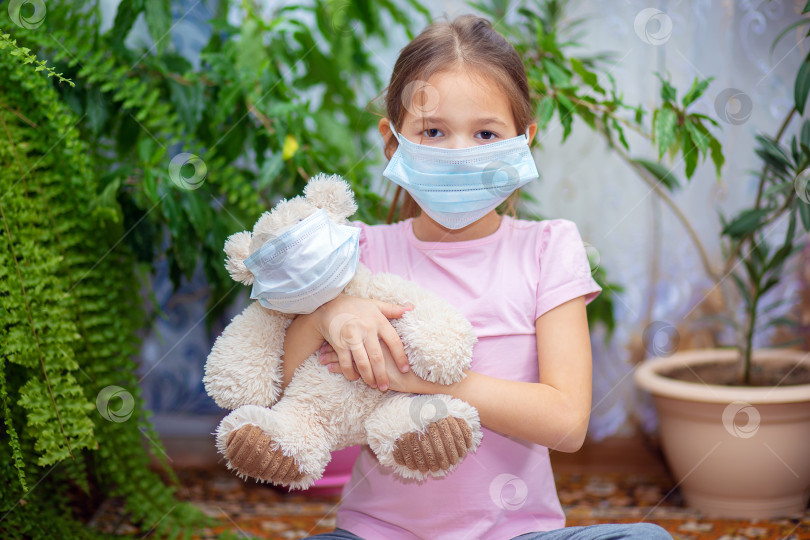 Скачать Девочка в медицинской маске сидит дома во время эпидемии во Вьесте со своим плюшевым мишкой, тоже в маске фотосток Ozero