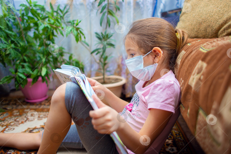 Скачать Девушка в медицинской маске сидит дома на полу и читает большую книгу. Проводить время дома. Детский досуг фотосток Ozero