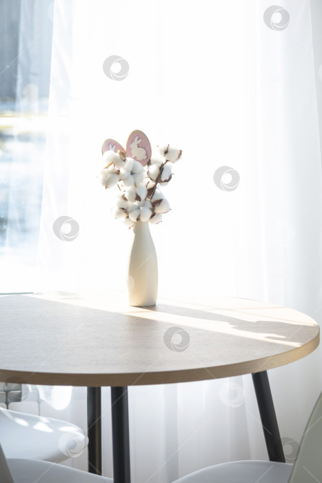 Скачать Букет из хлопковых веточек в вазе на круглом столе с пасхальным декором из яиц и пасхального кролика в свете из окна в минималистичном интерьере. фотосток Ozero