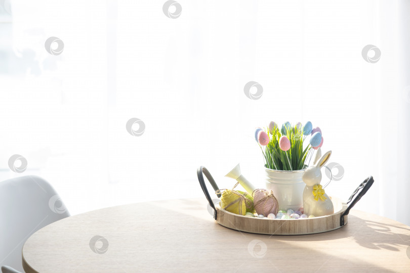 Скачать Пасхальное украшение из разноцветных яиц, свежая трава в лейке, пасхальный кролик на подносе на круглом столе в деревенском стиле. Праздничный интерьер загородного дома фотосток Ozero