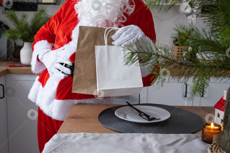 Скачать Санта-Клаус ставит его на кухонный стол и забирает бумажные пакеты с подарками для рукоделия, домашней выпечкой и доставкой еды. Покупки, переработка упаковки, ручная работа, доставка на Рождество и Новый год фотосток Ozero