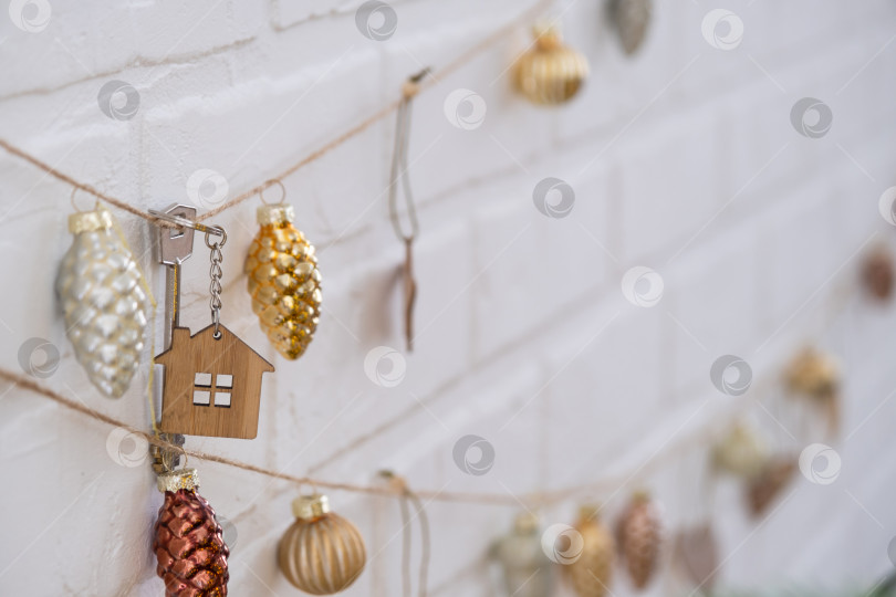 Скачать Ключ от дома с брелоком висит на гирлянде из елочных игрушек на стене. Подарок на Новый год, Рождество. Строительство, дизайн, проект, переезд в новый дом, ипотека, аренда и покупка недвижимости. фотосток Ozero