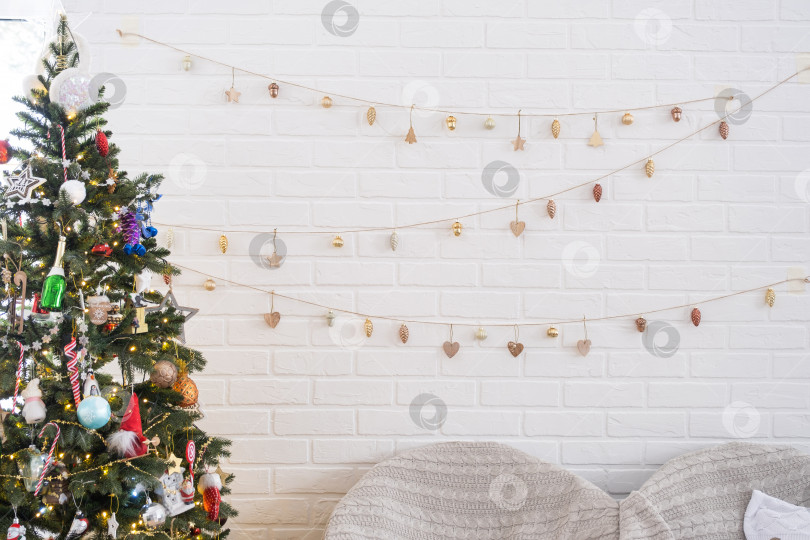 Скачать Рождественская елка в белом интерьере дома с кирпичными стенами в стиле лофт с гирляндами стеклянных игрушек на веревке. Светящиеся сказочные огни украшают комнату-студию. Уютный дом на Рождество и новый год фотосток Ozero