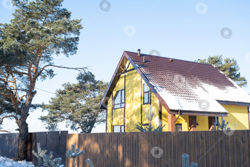 Скачать Желтый уютный домик в снегу зимой в деревне окружен соснами. Заснеженная крыша, трубы отопления и вентиляции, трапециевидные окна фотосток Ozero