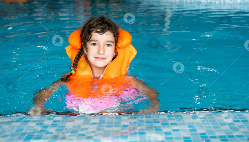 Скачать Ребенок в оранжевом спасательном жилете плавает в бассейне в аквакомплексе для семейного отдыха. Безопасность на воде, обучение плаванию фотосток Ozero