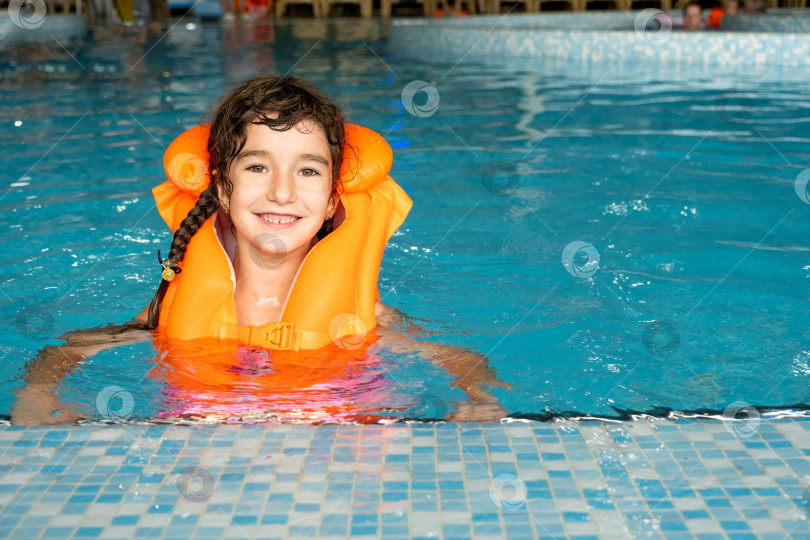 Скачать Ребенок в оранжевом спасательном жилете плавает в бассейне в аквакомплексе для семейного отдыха. Безопасность на воде, обучение плаванию фотосток Ozero