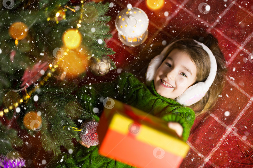 Скачать Девушка в теплом вязаном свитере и меховых наушниках лежит на одеяле возле рождественской елки с подарочной коробкой. Канун Рождества, мечта и желание. Новый год, праздничная атмосфера, гирлянды и боке фотосток Ozero