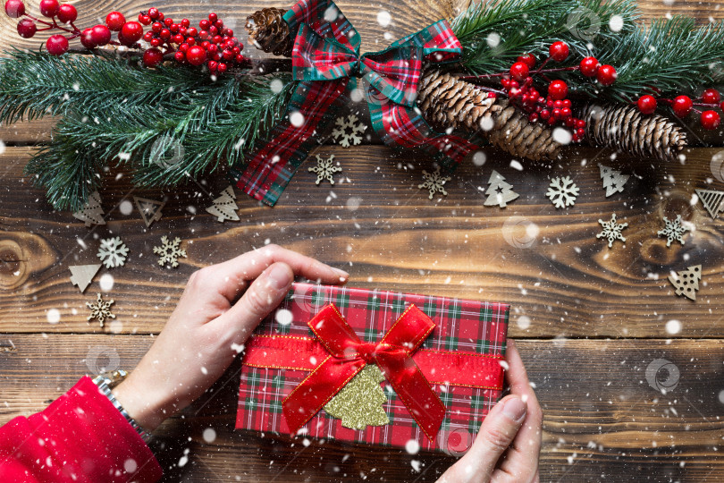 Скачать Руки, держащие подарочную коробку на деревянном рождественском фоне. Новый год, праздничное настроение, рождественская елка, снежинки, шишки. копировальное пространство, экологически чистое, ровное расположение. Праздничная упаковка фотосток Ozero