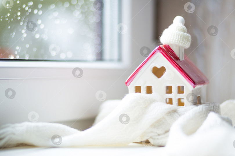 Скачать Уютный домик, закутанный в шапку и шарф в метель - декор подоконника. Зима, снег - утепление дома, защита от холода и непогоды, система отопления помещения. Праздничное настроение, Рождество, Новый год фотосток Ozero