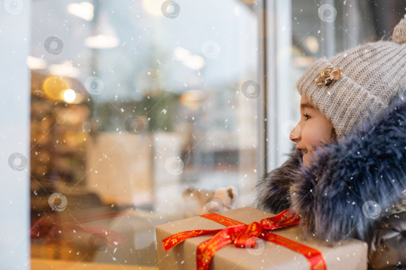 Скачать Портрет радостной девушки с подарочной коробкой на Рождество возле стеклянной витрины магазина зимой со снегом на праздничном рынке с украшениями и огнями. Теплая одежда, вязаная шапка, шарф и мех. Пространство для копирования фотосток Ozero