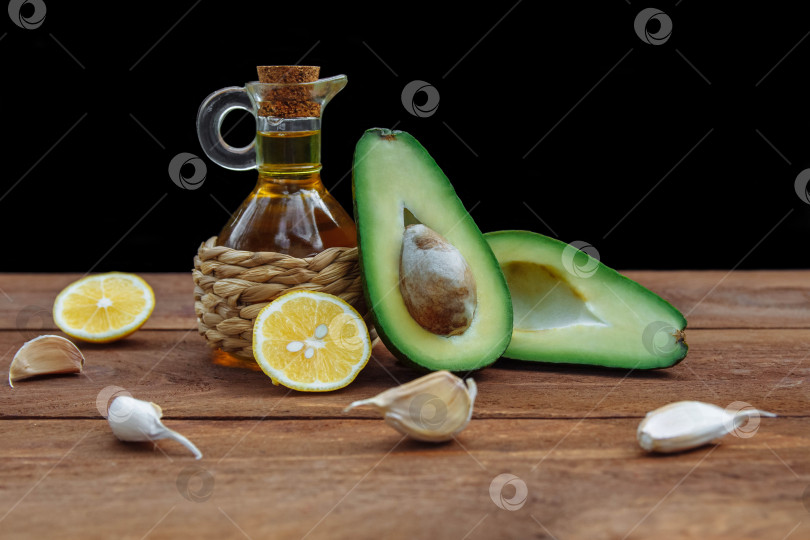 Скачать Зеленый авокадо, разрезанный пополам с косточкой, и стеклянная бутылка масла в плетеном футляре с ломтиками лимона и чесноком фотосток Ozero