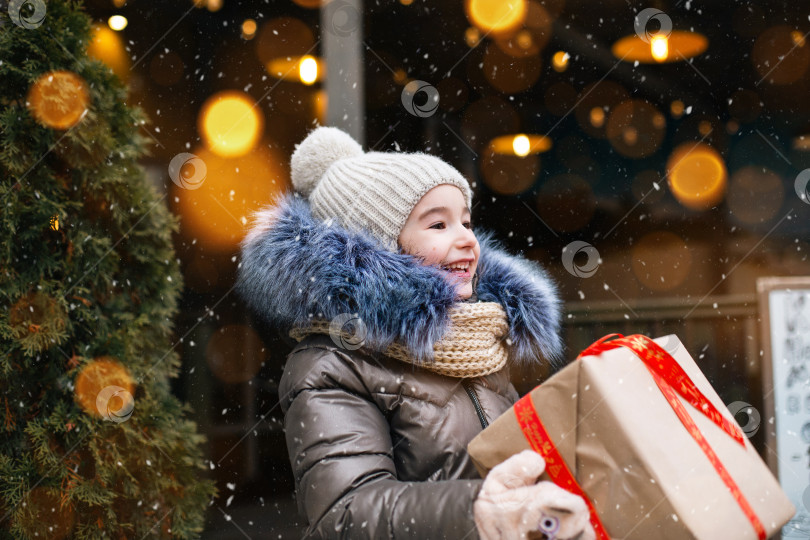 Скачать Портрет радостной девушки с подарочной коробкой на Рождество на городской улице зимой со снегом на праздничном рынке с украшениями и огнями. Теплая одежда, вязаная шапка, шарф и мех. Пространство для копирования фотосток Ozero