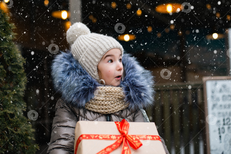 Скачать Портрет удивленной девушки с подарочной коробкой на Рождество на городской улице зимой со снегом на праздничном рынке с украшениями и огнями. Теплая одежда, вязаная шапка, шарф и мех. Пространство для копирования фотосток Ozero