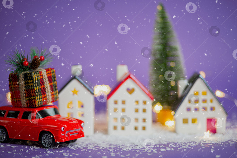 Скачать Рождественский декор - красный ретро-автомобиль на снегу проносится мимо домов с гирляндами в боке, рождественская елка с подарочными коробками на крыше. Игрушка на фиолетовом фоне. Новогодняя поздравительная открытка. Уютный дом фотосток Ozero