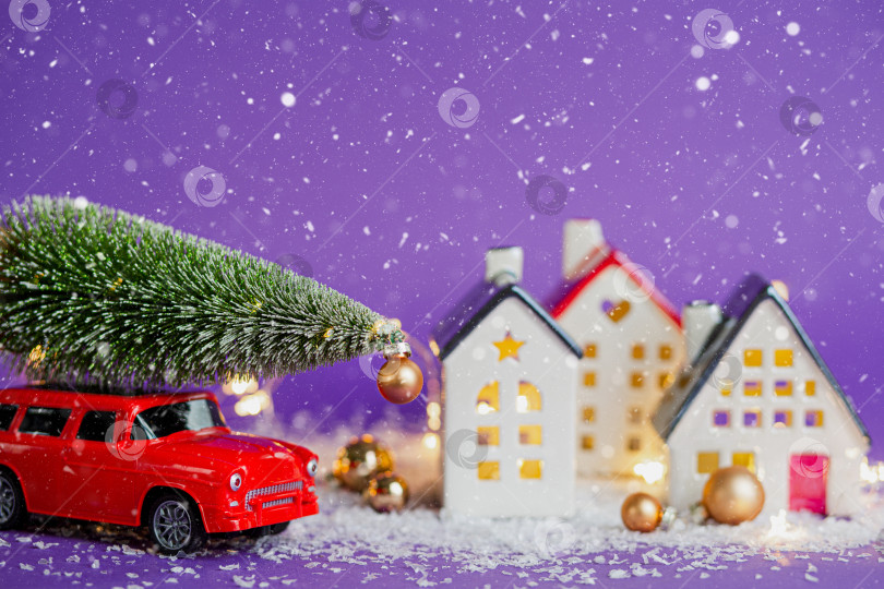 Скачать Рождественский декор - красный ретро-автомобиль на снегу проезжает мимо домов с гирляндами в боке, рождественская елка с подарочными коробками на крыше. Игрушка на фиолетовом фоне. Новогодняя поздравительная открытка. Уютный дом фотосток Ozero