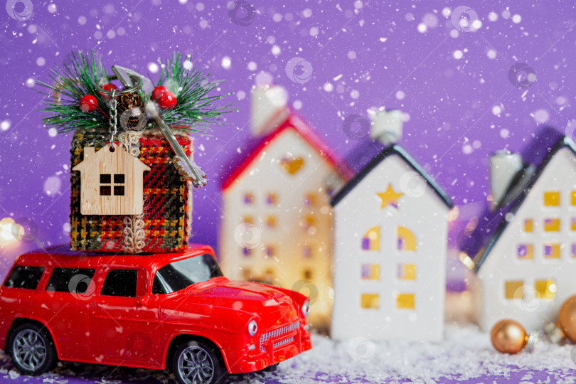 Скачать Красный ретро-автомобиль на снегу везет подарочную коробку с ключом от дома с брелком коттедж на крыше мимо домов с гирляндами, рождественская елка. Фиолетовый фон. Уютный Новый год. Недвижимость, переезд, ипотека. фотосток Ozero