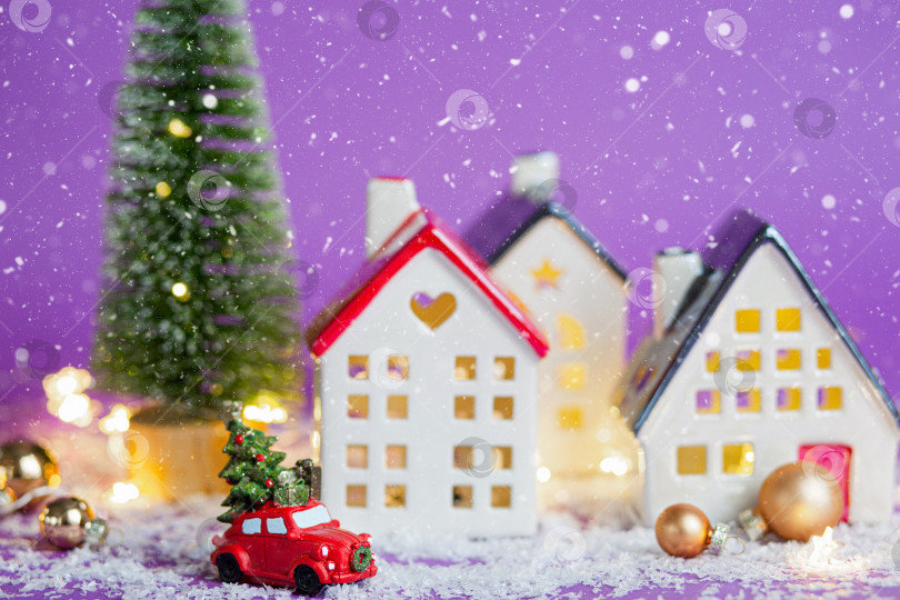 Скачать Рождественский декор - красный ретро-автомобиль на снегу проносится мимо домов с гирляндами в боке, рождественская елка с подарочными коробками на крыше. Игрушка на фиолетовом фоне. Новогодняя поздравительная открытка. Уютный дом фотосток Ozero