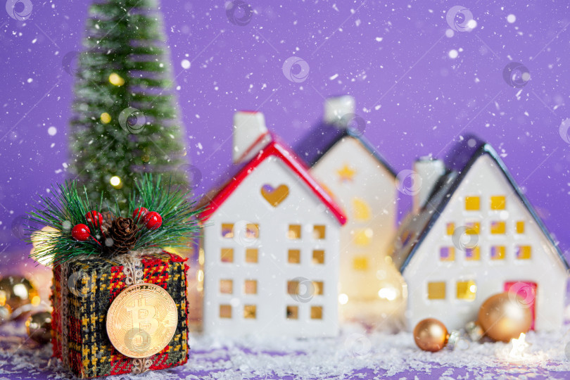 Скачать Биткоин-монета на подарочной коробке мимо домов с гирляндами и снегом, рождественской елки. Фиолетовый фон очень красивый. Новогодняя поздравительная открытка. Криптовалюта, финансы, богатство и инвестирование. фотосток Ozero