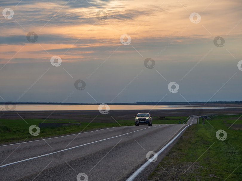 Скачать Автомобиль едет по дороге в сельской местности среди природы на закате. Асфальтированная дорога рядом с заливом морской лагуны фотосток Ozero