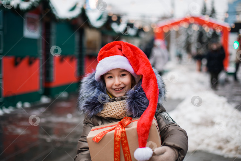 Скачать Портрет радостной девушки в шляпе Санты с подарочной коробкой на Рождество на улице города зимой со снегом на праздничном рынке с украшениями и сказочными огнями. Теплая одежда, вязаный шарф и мех. Новый год фотосток Ozero