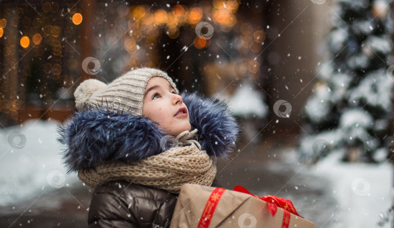 Скачать Счастливая девочка с подарочной коробкой на Рождество ловит языком снежинки в зимнем городе со снегом на праздничном рынке с украшениями и огнями. Теплая одежда, вязаная шапка, шарф и мех. фотосток Ozero