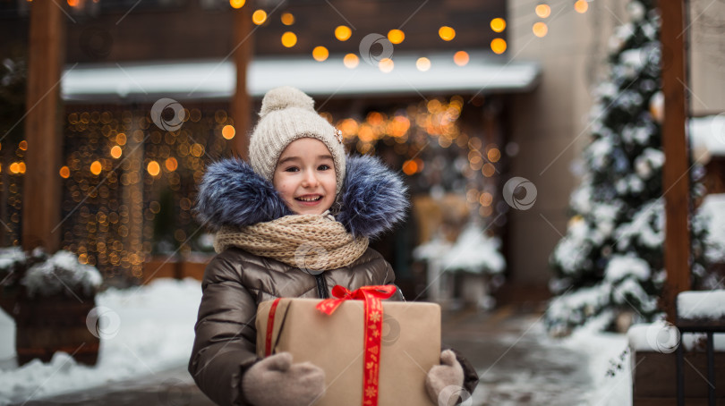 Скачать Портрет радостной девушки с подарочной коробкой на Рождество на городской улице зимой со снегом на праздничном рынке с украшениями и гирляндами. Теплая одежда, вязаная шапка, шарф и мех. Новый год фотосток Ozero