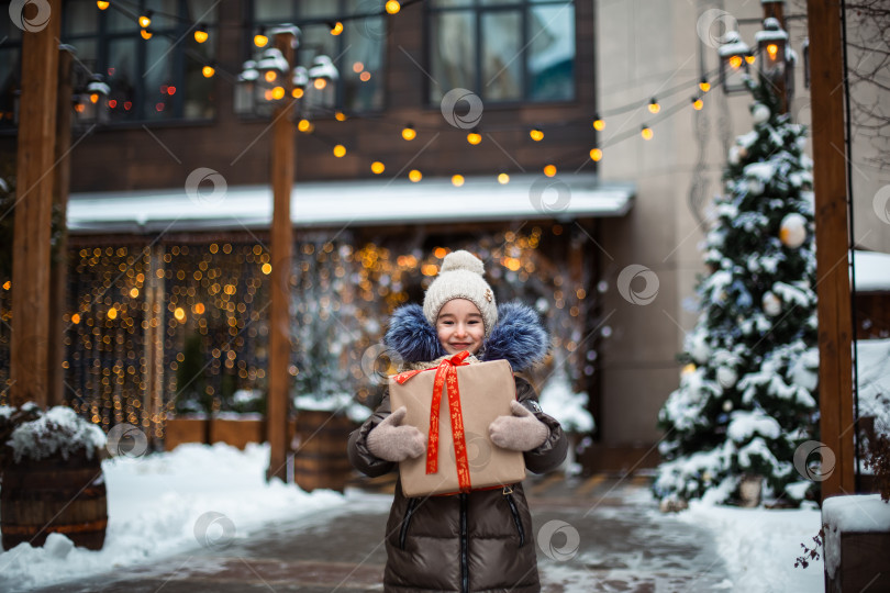 Скачать Портрет радостной девушки с подарочной коробкой на Рождество на городской улице зимой со снегом на праздничном рынке с украшениями и гирляндами. Теплая одежда, вязаная шапка, шарф и мех. Новый год фотосток Ozero