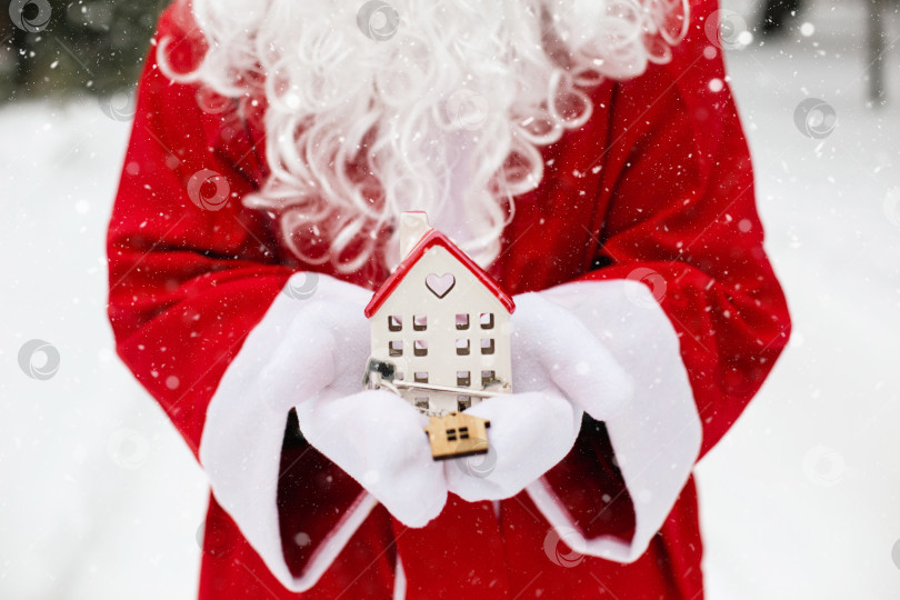 Скачать Ключ от дома с брелком-коттеджем в руках Санта-Клауса на открытом воздухе в снегу. Сделки с недвижимостью, покупка, строительство, переезд, ипотека. Уютный дом. Счастливого Рождества, новогоднего бронирования мероприятия и зала фотосток Ozero