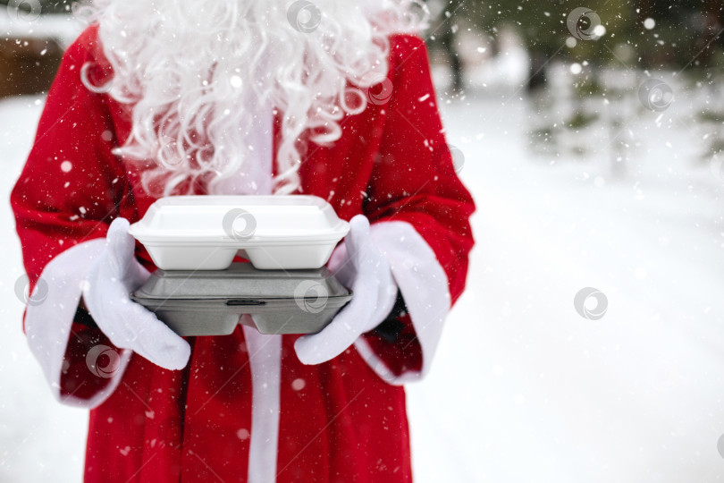 Скачать Контейнеры службы доставки еды в руках Санта-Клауса на открытом воздухе в снегу. Акция в канун Рождества. Готовый горячий заказ, одноразовая пластиковая коробка. Организация питания на новогодние праздники. Место для копирования, макет фотосток Ozero
