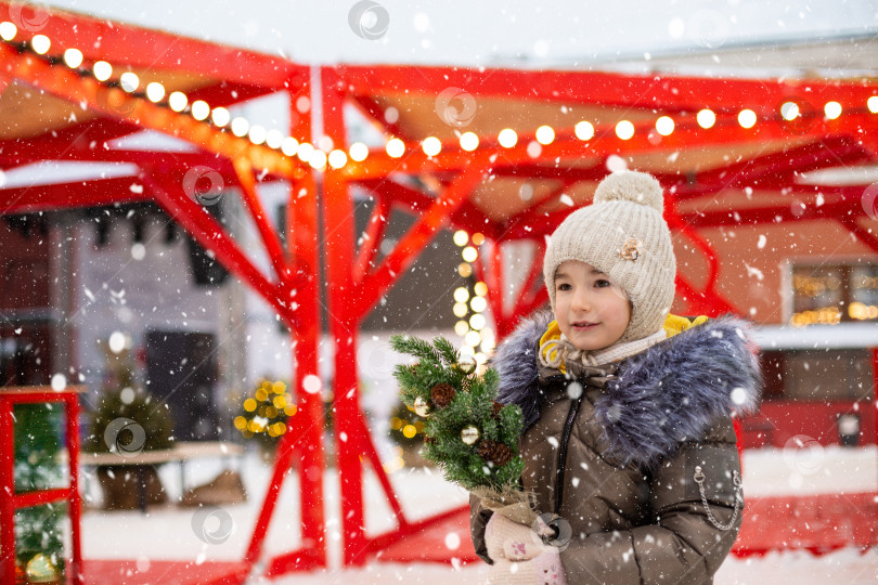 Скачать Девушка с рождественской елкой в руках на открытом воздухе в теплой одежде зимой на праздничном рынке. Сказочные огни и гирлянды украсили снежный городок к новому году фотосток Ozero