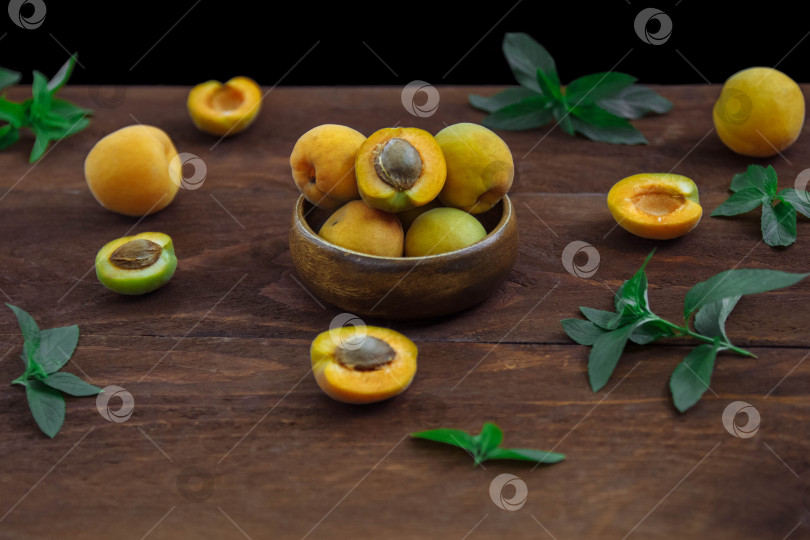 Скачать Спелые, сочные, сладкие желтые абрикосы выложите в миску целиком и разрежьте пополам на коричневом деревянном столе с зелеными листьями мяты фотосток Ozero