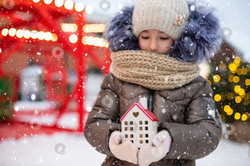 Скачать Дом, миниатюрный коттедж в руках девочки, одетой в варежки и теплую одежду на улице в снегу. Семейные ценности, покупка жилья, переезд, ипотека. Бронирование уютного дома на Рождество, новый год фотосток Ozero