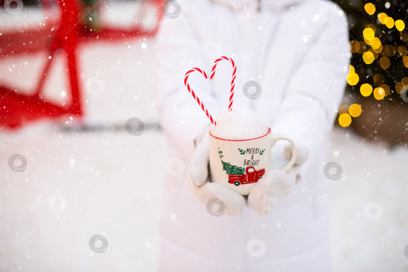 Скачать Женщина с кружкой со снегом, леденцовой палочкой и надписью Merry and Bright в руках на улице в теплой одежде на зимнем праздничном рынке. Гирлянды сказочных огней украсили снежный городок к новому году. Рождество фотосток Ozero