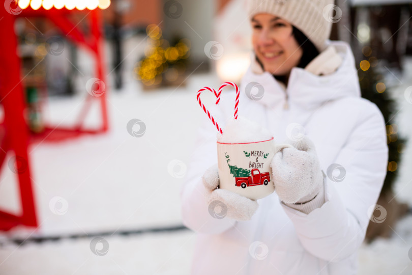 Скачать Женщина с кружкой со снегом, леденцовой палочкой и надписью Merry and Bright в руках на улице в теплой одежде на зимнем праздничном рынке. Гирлянды сказочных огней украсили снежный городок к новому году. Рождество фотосток Ozero