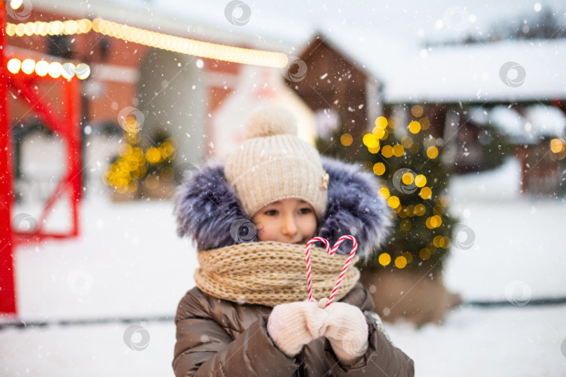 Скачать Симпатичная девушка держит в руках леденцовый тростник в форме сердца на открытом воздухе в теплой одежде на зимнем праздничном рынке. Гирлянды сказочных огней украсили снежный городок к новому году. Рождественское настроение фотосток Ozero