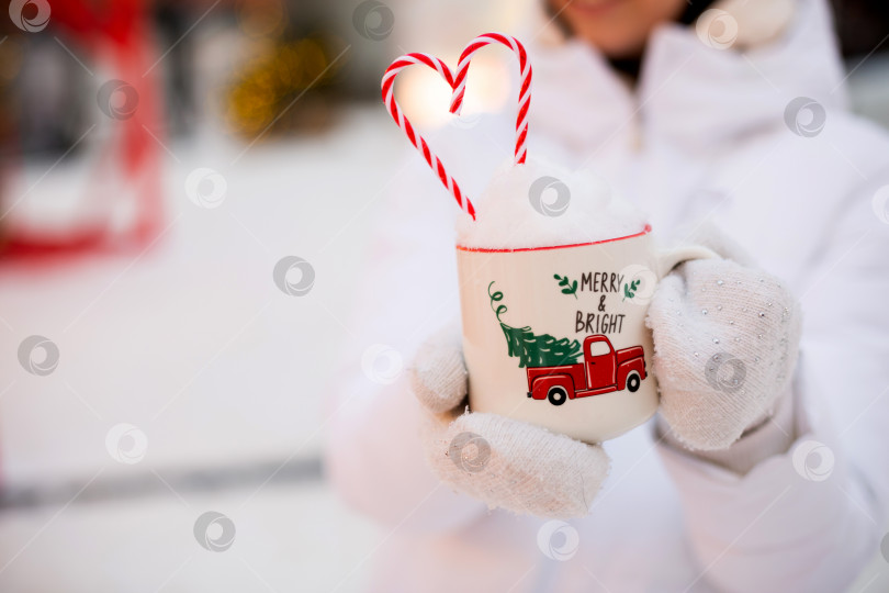 Скачать Женщина с кружкой со снегом, леденцовой палочкой и надписью Merry and Bright в руках на улице в теплой одежде на зимнем праздничном рынке. Гирлянды сказочных огней украсили снежный городок к новому году. Рождество фотосток Ozero