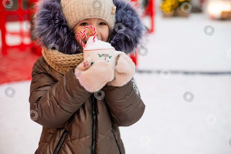 Скачать Девушка с кружкой со снегом, леденцовой палочкой и надписью Merry and Bright в руках на улице в теплой одежде зимой на праздничном рынке. Гирлянды сказочных огней украсили снежный городок к новому году. Рождество фотосток Ozero