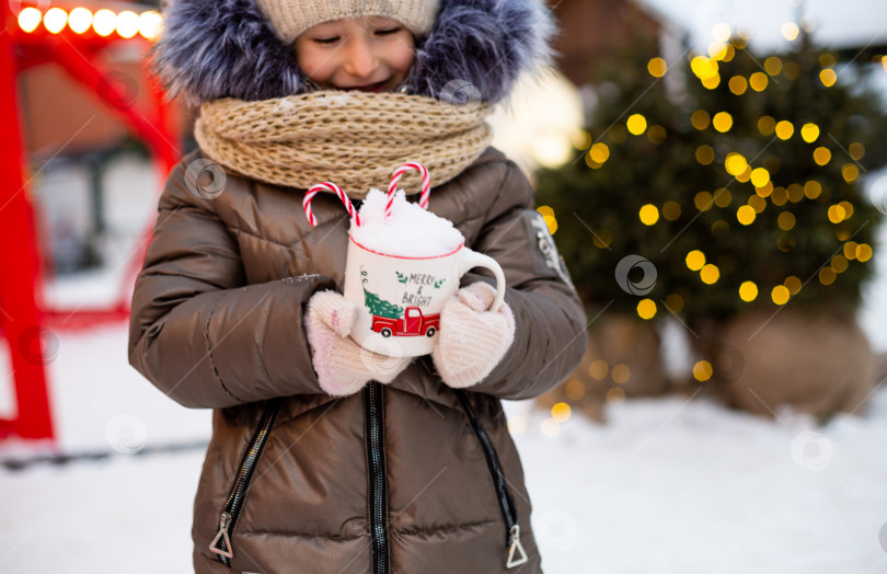 Скачать Девушка с кружкой со снегом, леденцовой палочкой и надписью Merry and Bright в руках на улице в теплой одежде зимой на праздничном рынке. Гирлянды сказочных огней украсили снежный городок к новому году. Рождество фотосток Ozero